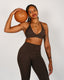 Woman in brown sports bra and leggings holding a leather medicine ball against a plain background